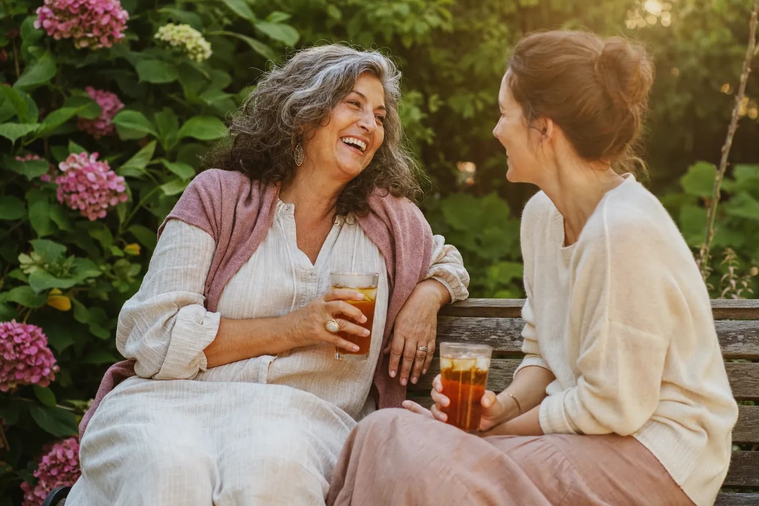 An aunt and her adult niece laughing together over iced tea on a garden bench