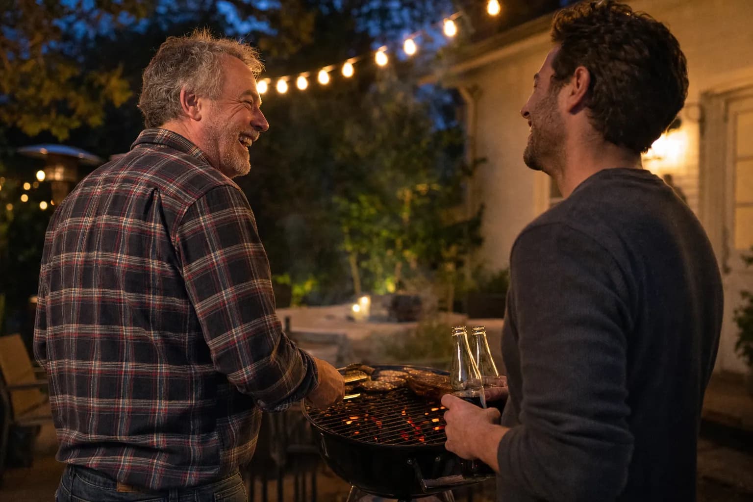 An uncle and his adult nephew laughing together at a backyard grill at dusk