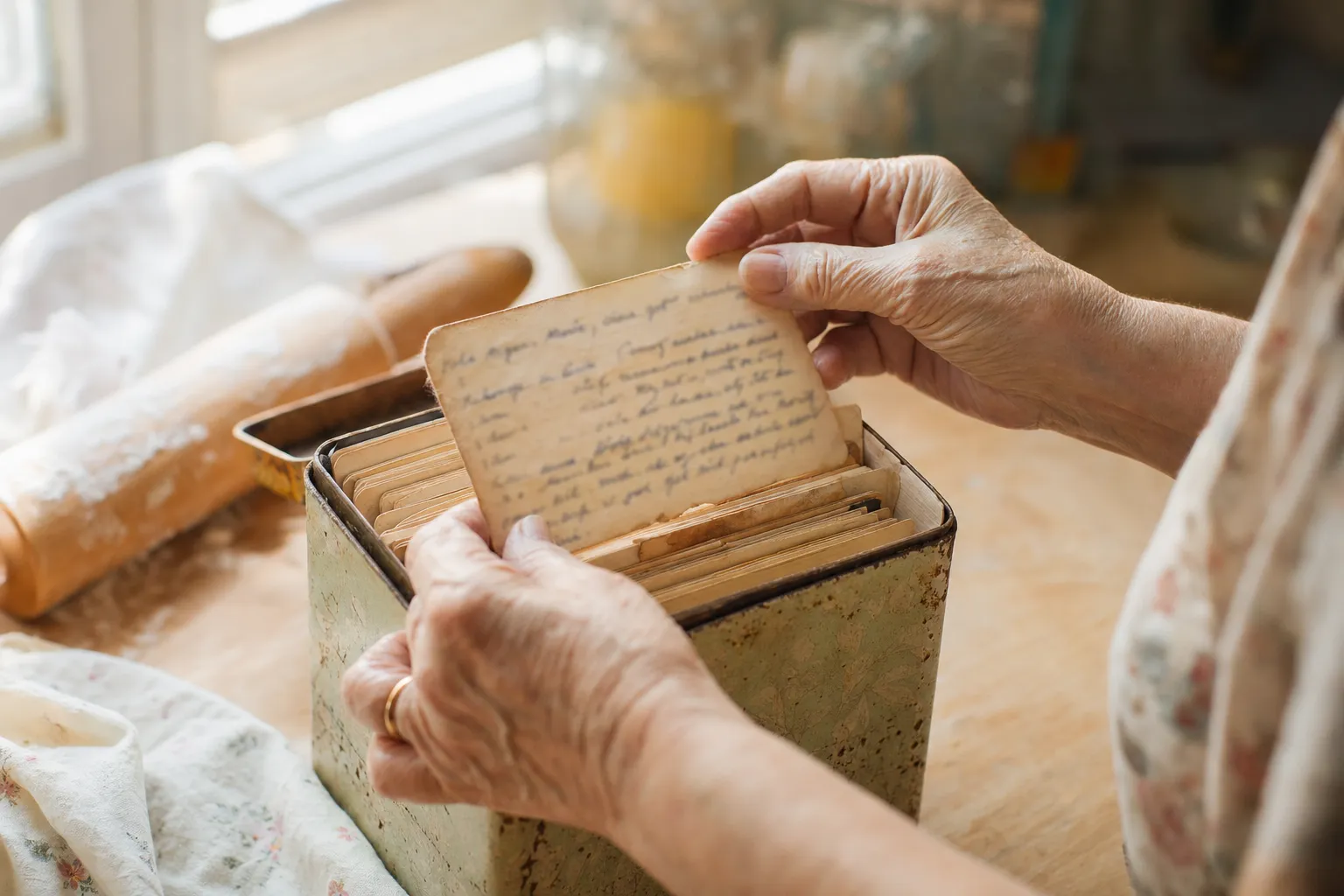 A mother's gentle hands lifting a worn handwritten recipe card from a vintage tin recipe box